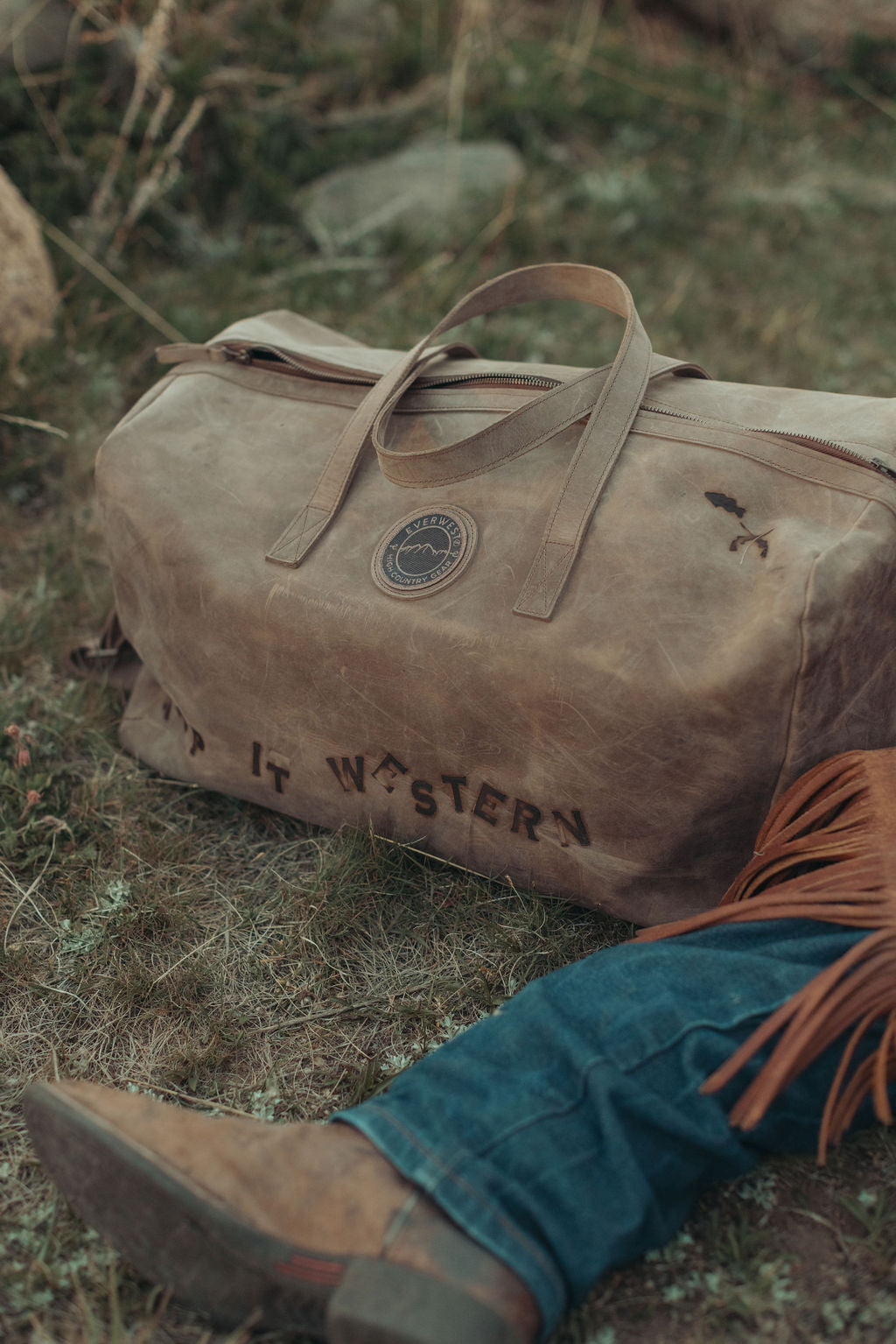 A distressed large brown leather duffel bag with 'KEEP IT WESTERN' custom branded on the side, resting on grassy ground next to a person's leg in jeans and a brown leather boot with orange fringe.