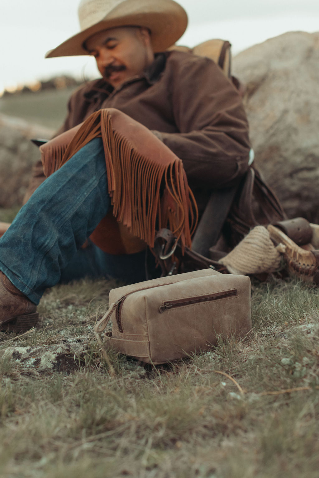 A man in a cowboy hat and brown jacket sits outdoors on grass beside large rocks, with a distressed brown leather toiletry kit placed in the foreground.