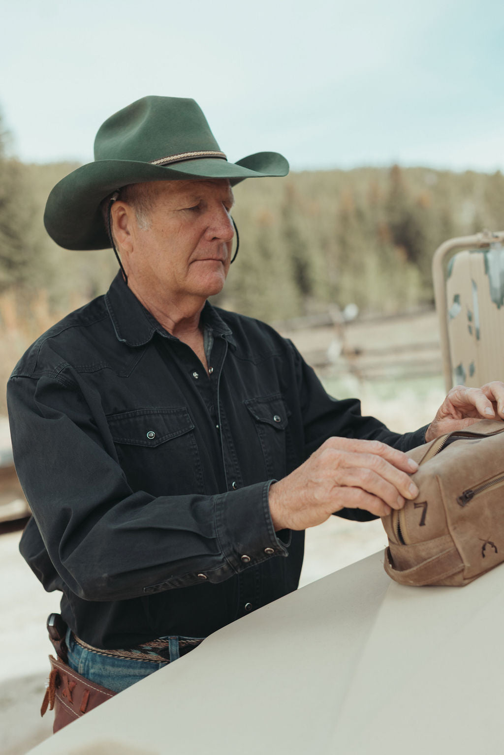 A man wearing a dark green cowboy hat, black long-sleeve shirt, and jeans sits outdoors at a table, holding a brown leather dopp kit with custom brands on it.
