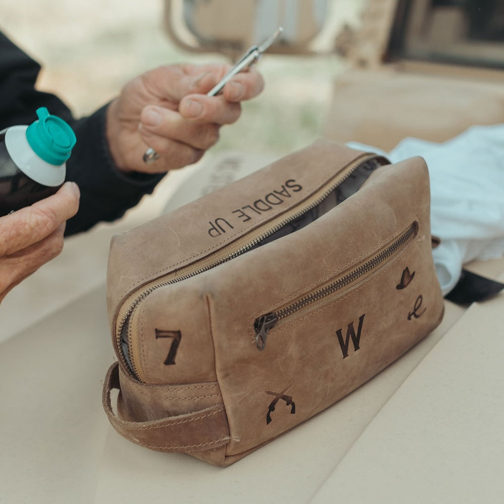A distressed brown leather dopp kit with custom branding letters and symbols, shown on a table with a person's hands holding a razor nearby.