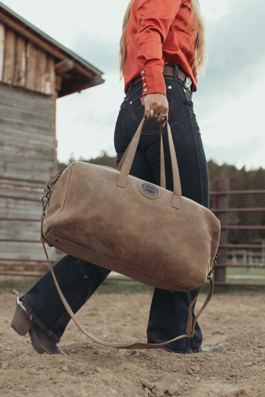 A woman in a sandy barn area carries a large distressed brown leather duffel bag with the Everwest logo patch stitched into the side of the bag.