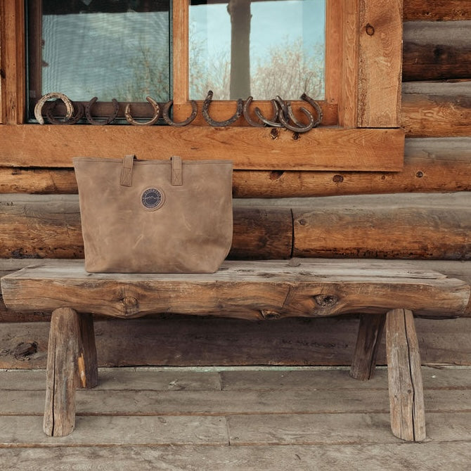 Brown leather tote bag resting on bench in front of a log cabin.