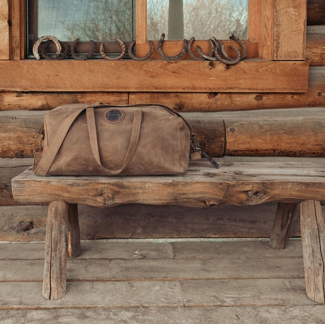 A brown leather duffel bag with leather handles sits on a rustic wooden bench in front of a log cabin wall with horseshoe decorations.