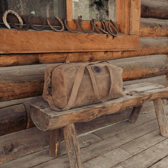 A brown leather duffel bag with dual shoulder straps and the Everwest logo patch, resting on a rustic wooden bench in front of a log cabin wall with horseshoes on the window.