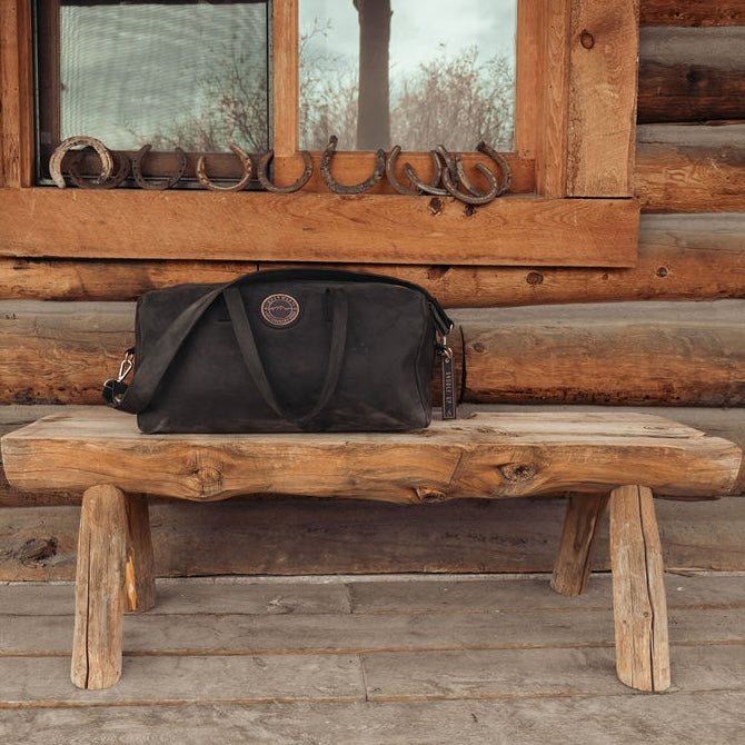 A black leather duffel bag with the Everwest logo patch sits on a rustic wooden log bench in front of a log cabin wall with horseshoe decorations.