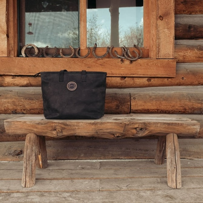 A black leather tote bag with the Everwest brand logo sits on a rustic wooden bench in front of a log cabin wall with a window and horseshoe decorations.