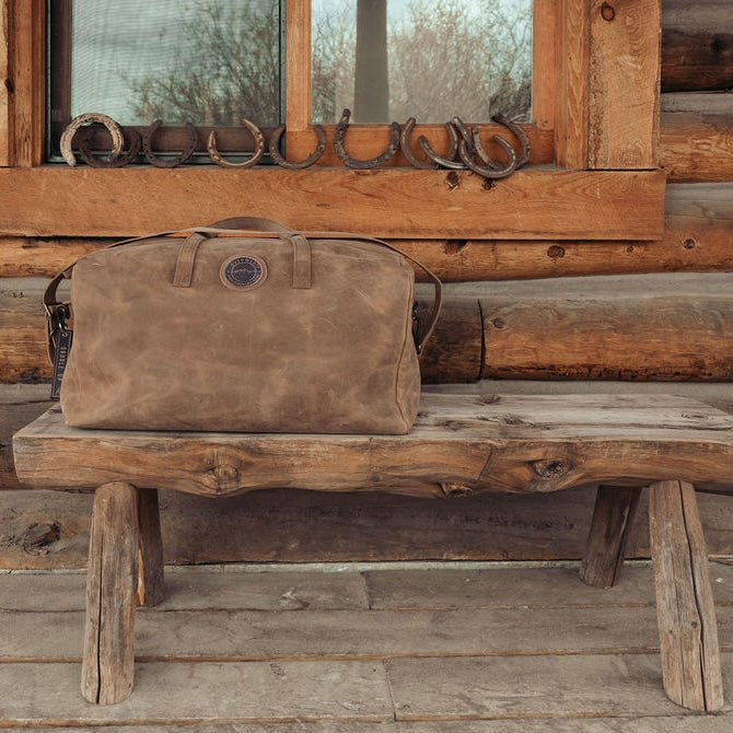 A brown leather duffle bag with the Everwest logo patch sits on a rustic wooden bench in front of a log cabin window.