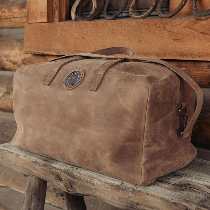 A large brown distressed leather duffel bag with the Everwest logo patch, resting on a rustic wooden bench against a wooden log wall background.