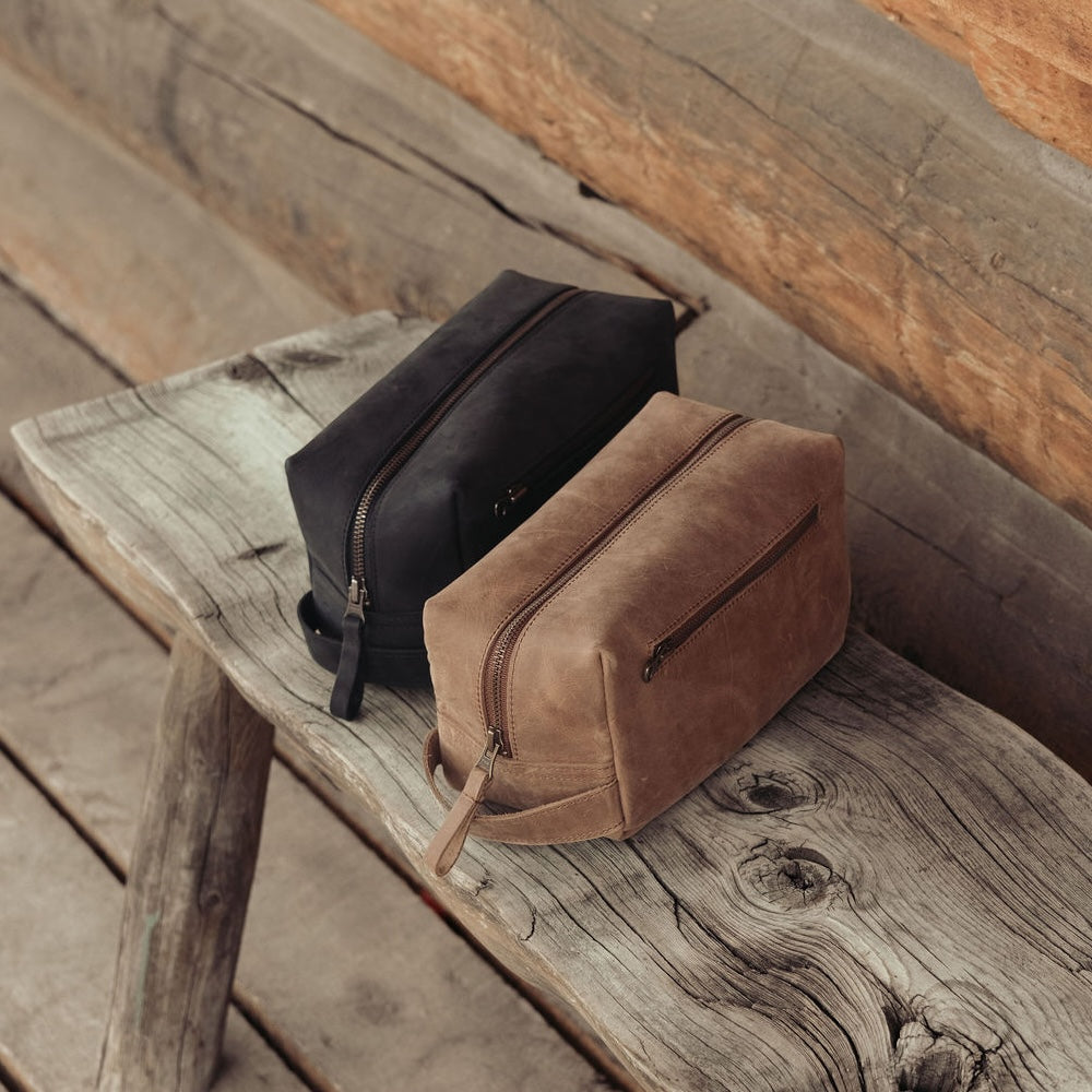 Two leather dopp kits, one black and one brown, placed on a rustic wooden bench in front of a log wall.