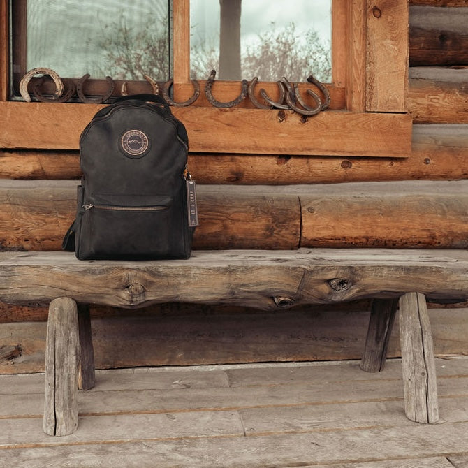 A black leather backpack with the Everwest logo patch sits on a rustic wooden log bench in front of a log cabin wall with horseshoes hanging on the window frame.
